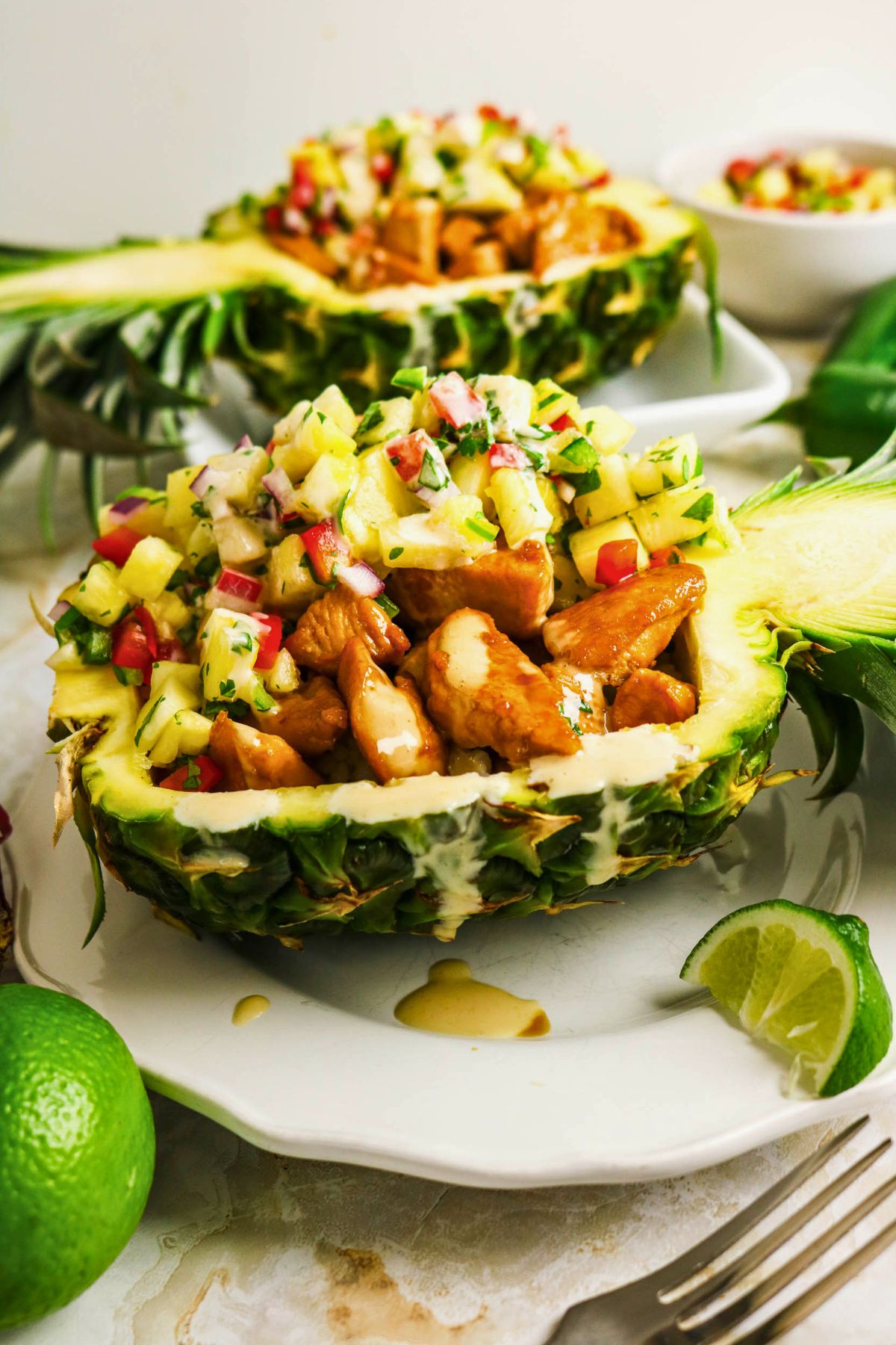 Pineapple Chicken Bowls: Halved pineapples filled with cooked chicken pieces and fresh pineapple salsa, garnished with herbs, on a white plate with limes on the side.