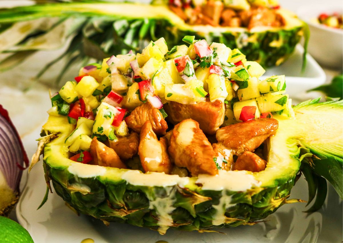 Pineapple Chicken Bowls feature juicy chicken pieces and vibrant fruit salsa served in a hollowed-out pineapple half, garnished with sauce and fresh herbs.