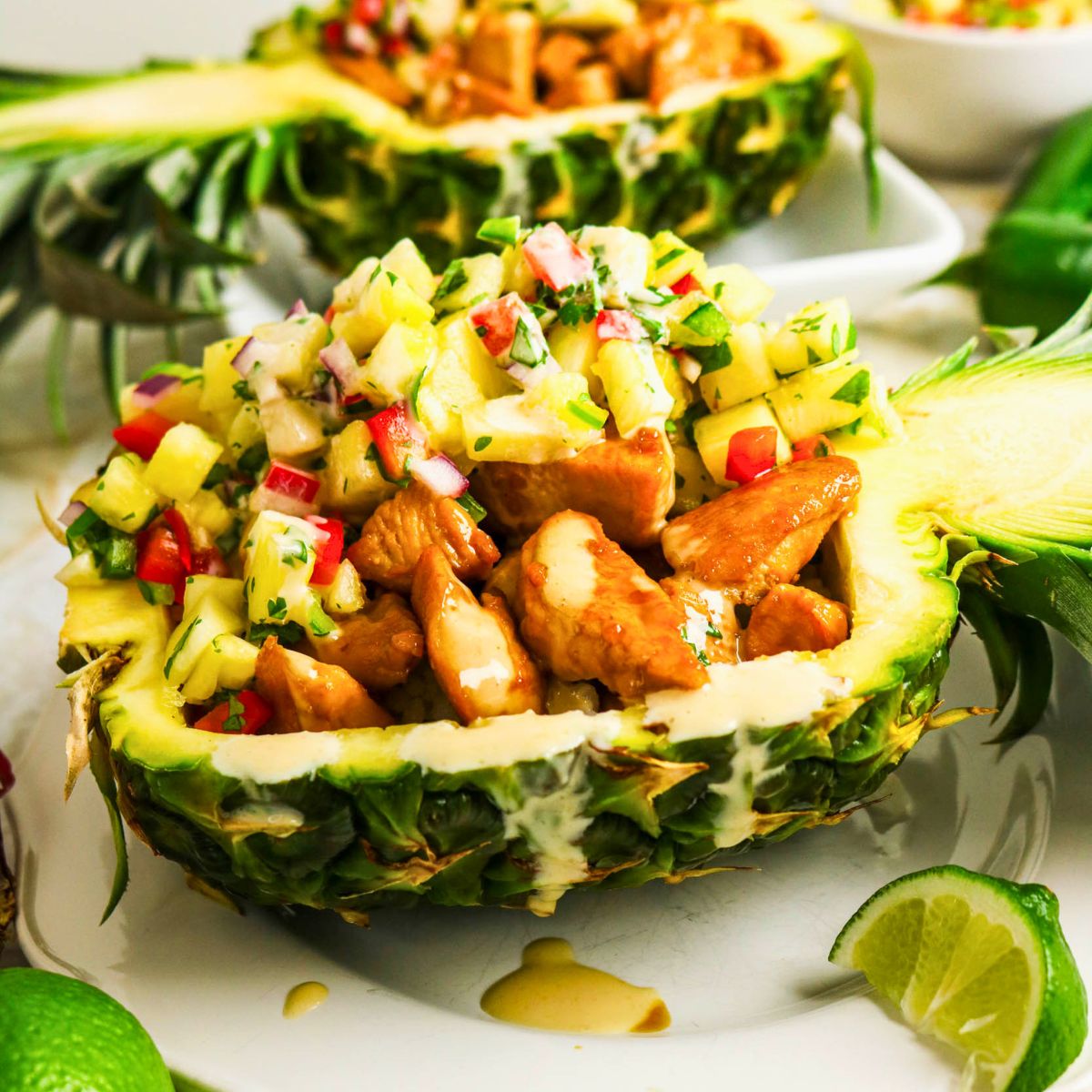 Enjoy vibrant Pineapple Chicken Bowls with juicy chicken and pineapple salsa served in a halved pineapple bowl, garnished with sauce and fresh lime wedges on the side.