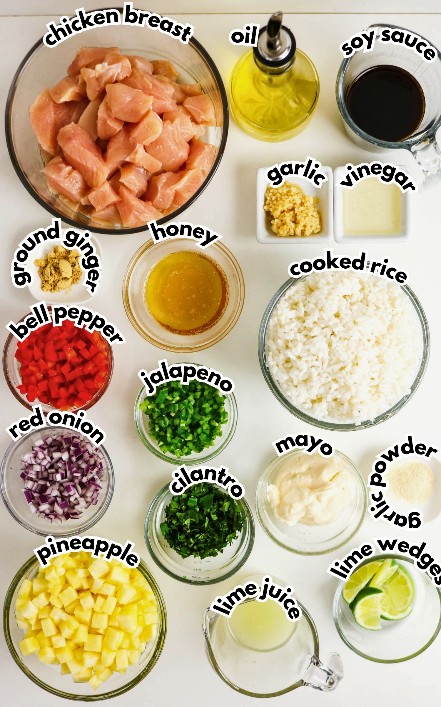 Overhead view of Pineapple Chicken Bowls ingredients—chopped chicken breast, cooked rice, diced vegetables, pineapple, sauces, spices, and condiments—arranged in labeled bowls on a white surface.