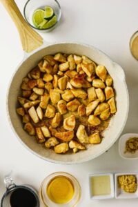 Overhead view of cooked chicken pieces in a skillet, surrounded by small bowls containing lime wedges, sauces, and seasonings on a white surface—perfect for assembling flavorful Pineapple Chicken Bowls.