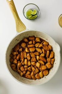 Cooked, cubed chicken pieces in a white skillet with brown sauce, perfect for Pineapple Chicken Bowls, placed on a white surface next to a small bowl of lime wedges.