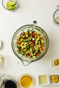A glass bowl filled with chopped vegetables and herbs sits on a white surface, surrounded by small bowls of ingredients—perfect for assembling fresh Pineapple Chicken Bowls with lime wedges and flavorful liquids.