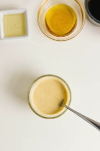A bowl of creamy dressing with a spoon, surrounded by small containers of oil, honey, and soy sauce on a white surface—perfect for drizzling over Pineapple Chicken Bowls.