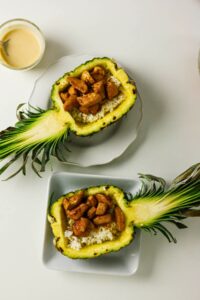 Two Pineapple Chicken Bowls feature pineapple halves filled with white rice and glazed chicken pieces, served on plates alongside a small bowl of sauce.