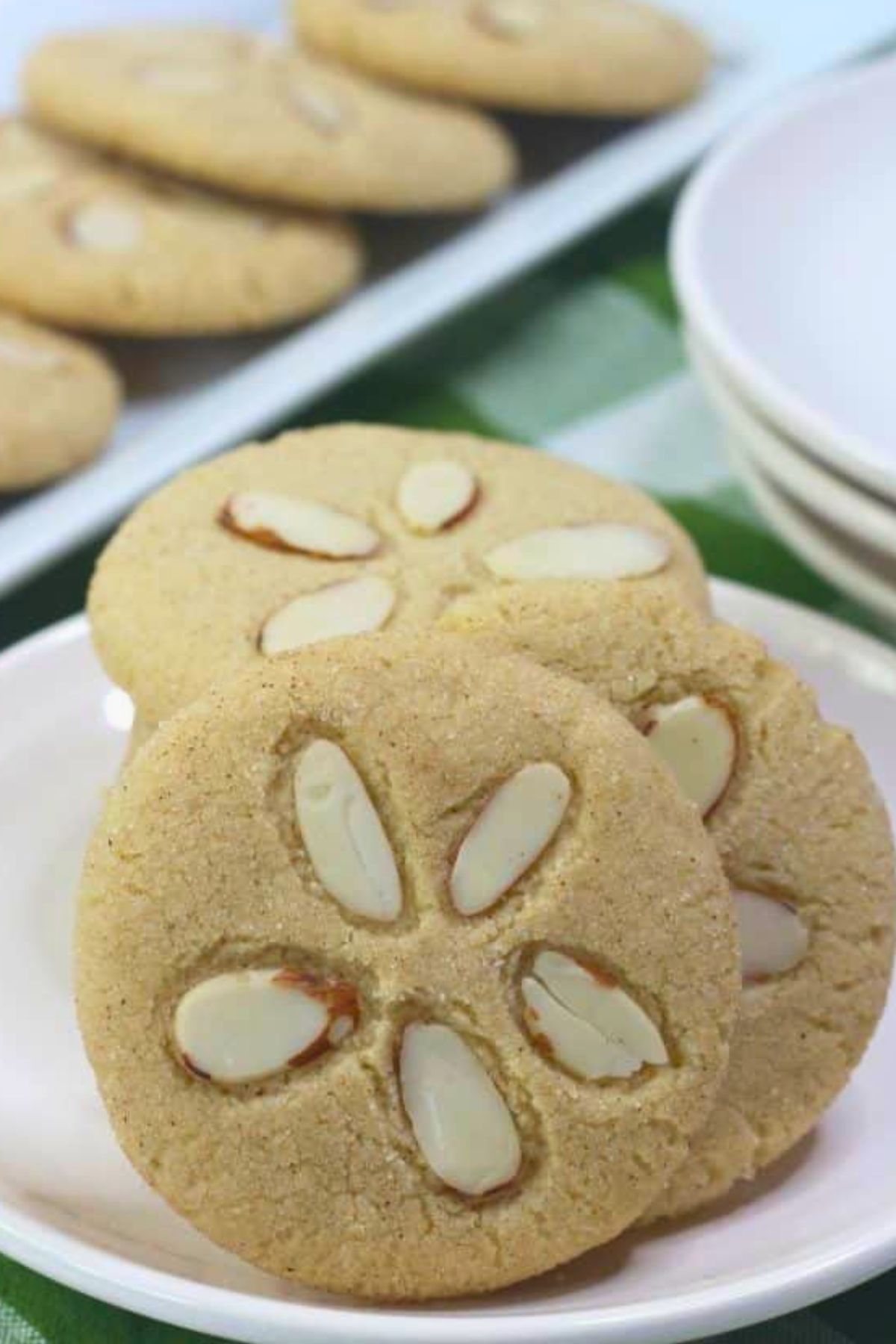 Two round cookies topped with sliced almonds are on a white plate, with more cookies and stacked plates visible in the background.