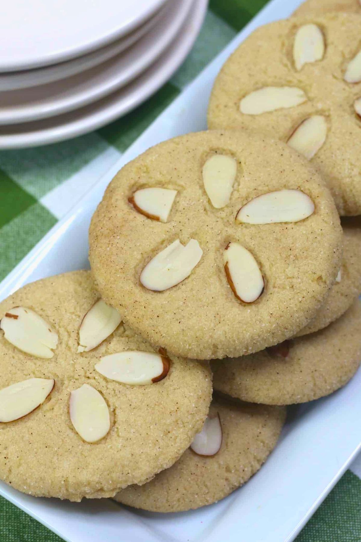 A plate of round almond cookies, each topped with slivered almonds, arranged on a white rectangular dish next to stacked plates.
