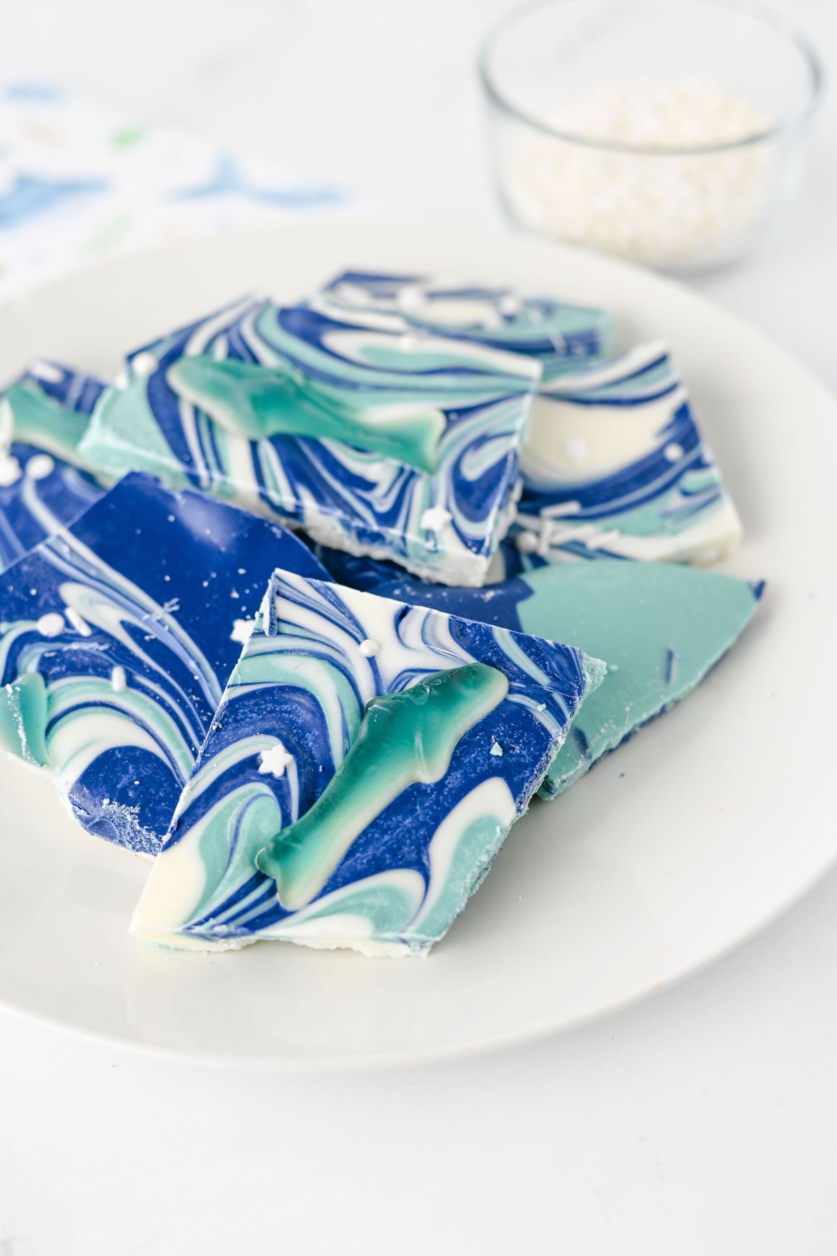 A plate with pieces of swirled blue, green, and white chocolate bark. A small bowl with white sprinkles is in the background.