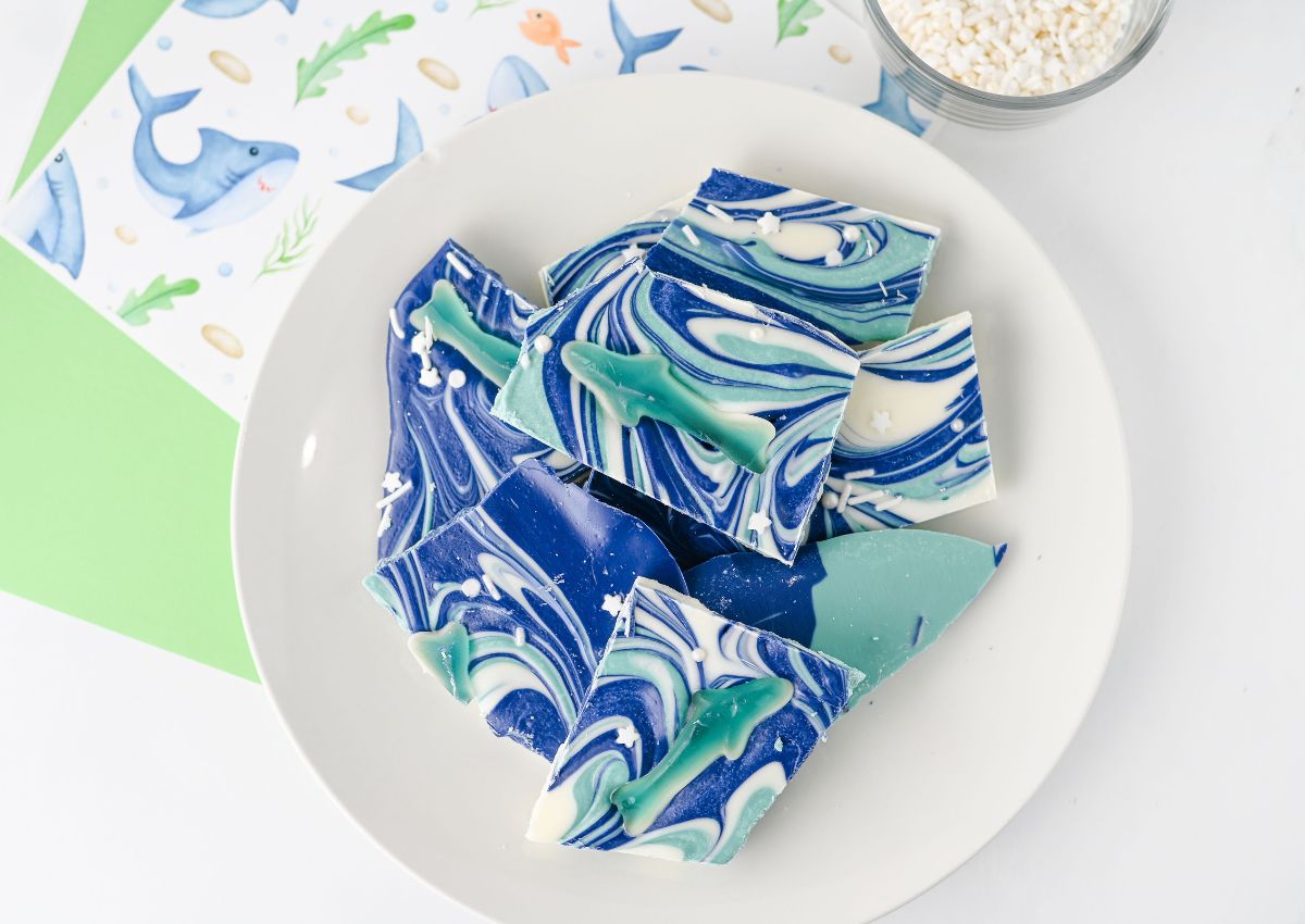 A white plate with pieces of blue, green, and white marbled chocolate bark, some shaped like sharks, sits on a white surface near decorative shark-themed paper.