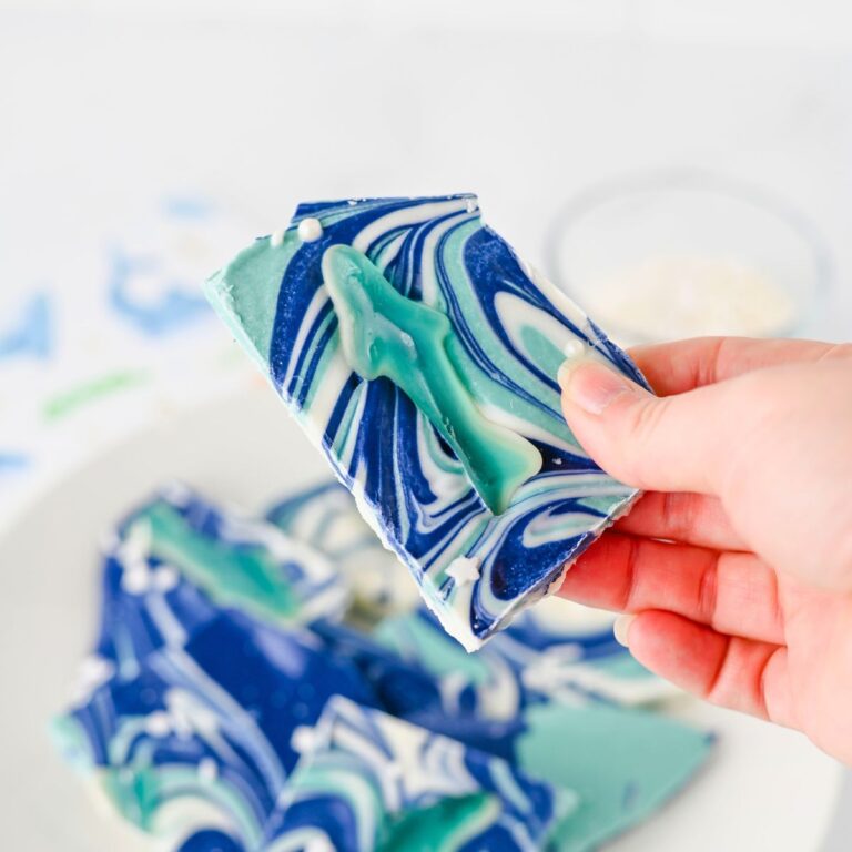 A hand holds a piece of blue and white swirled chocolate bark topped with a gummy fish; more pieces are visible on a white plate in the background.