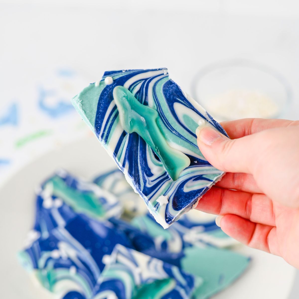 A hand holds a piece of blue and white swirled chocolate bark topped with a gummy fish; more pieces are visible on a white plate in the background.