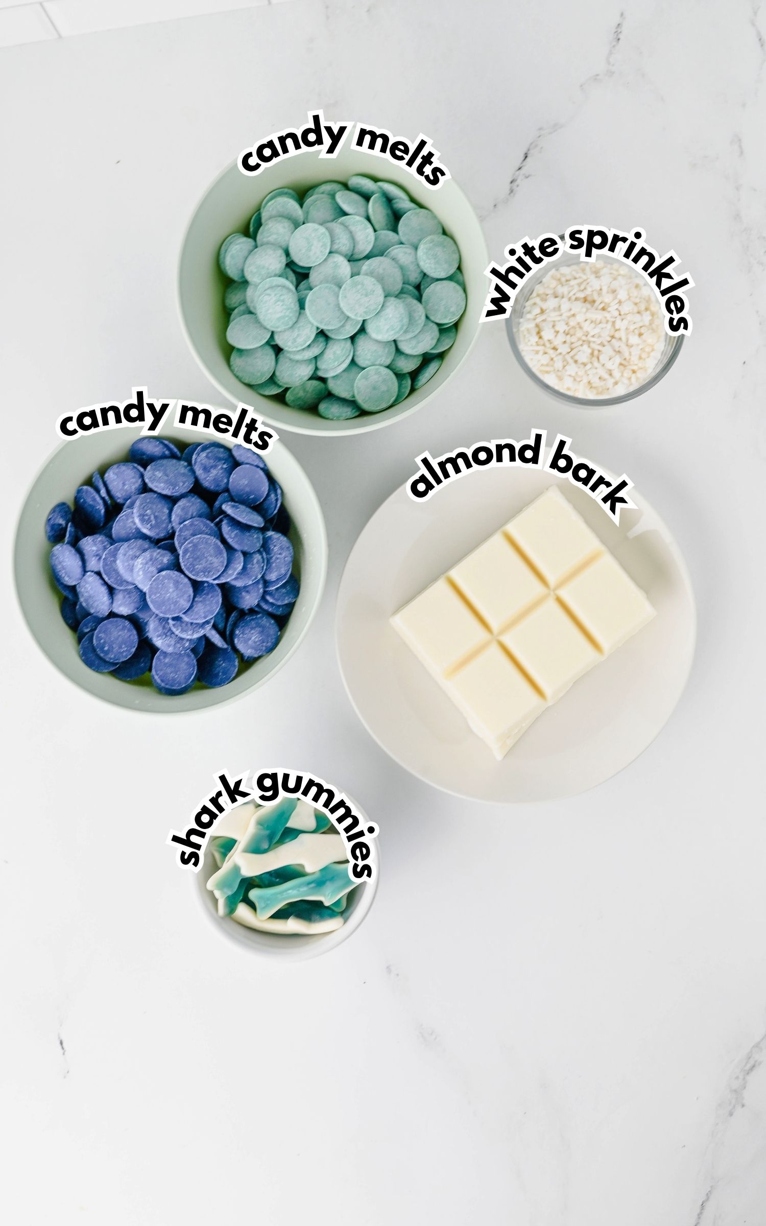 Four bowls containing blue candy melts, teal candy melts, white sprinkles, almond bark squares, and blue shark gummies arranged on a white surface.