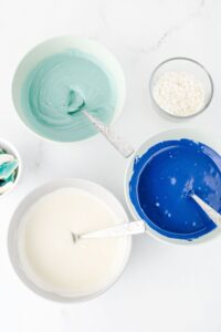 Three bowls with blue, teal, and white melted candy coating and spoons; a small bowl of white sprinkles sits nearby on a white surface.