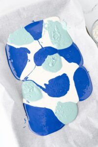 Melted blue, light blue, and white chocolate are spread in irregular patches on parchment paper, ready to be mixed or set.