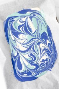 A rectangular sheet of marbled blue, teal, and white chocolate rests on parchment paper.