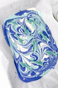 A sheet of blue, white, and teal swirled chocolate bark topped with gummy sharks and white star sprinkles on parchment paper.