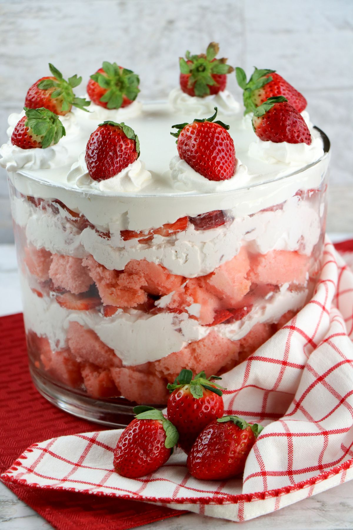 A trifle dessert with layers of whipped cream, strawberry cake, and sliced strawberries in a glass bowl, topped with whole strawberries.