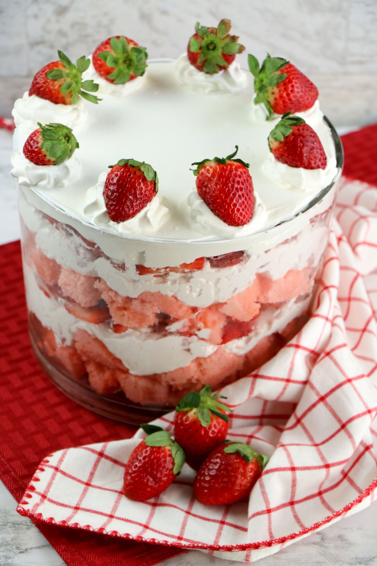 A layered strawberry trifle with whipped cream in a glass bowl, topped with whole strawberries, sits on a red and white checkered cloth with strawberries beside it.