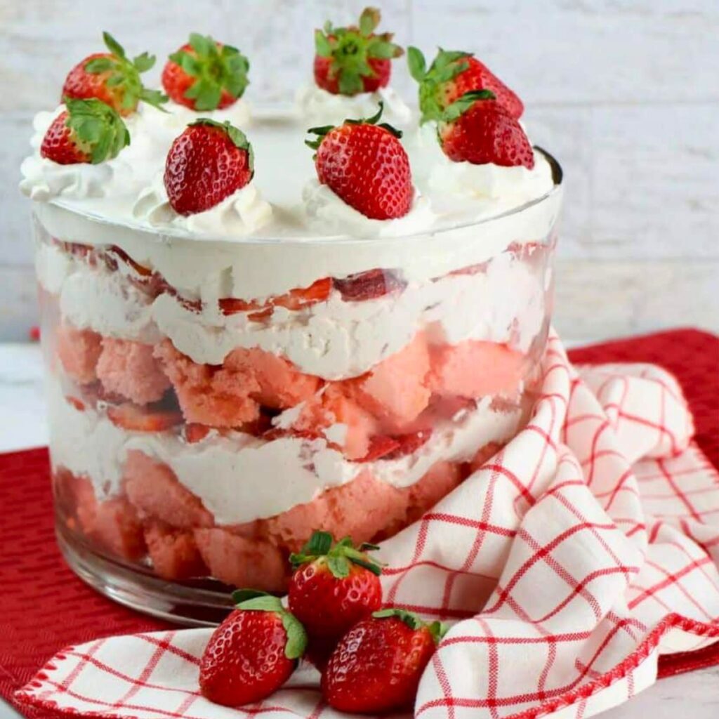 A glass trifle bowl filled with layers of whipped cream, strawberry pieces, and pink cake, topped with whole strawberries, sits on a red and white checkered cloth.