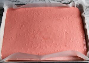 A rectangular baking tray lined with parchment paper holds a layer of pink cake batter with a slightly bubbly surface.