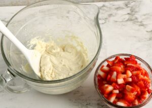 A glass bowl with creamy batter and a spatula next to a smaller bowl filled with chopped strawberries on a white countertop.