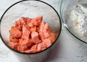 A glass bowl filled with chunks of pink cake next to a bowl containing white whipped cream or frosting on a white surface.