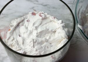 A glass bowl filled with a whipped cream mixture that contains small pieces of fruit, set on a marble surface.
