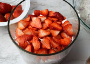 Sliced strawberries layered on top of a dessert in a glass bowl, with more strawberries and another dish visible in the background.