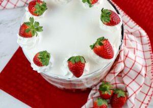 A glass trifle dish filled with a dessert topped with white whipped cream and whole strawberries, set on a red and white checkered cloth with extra strawberries nearby.