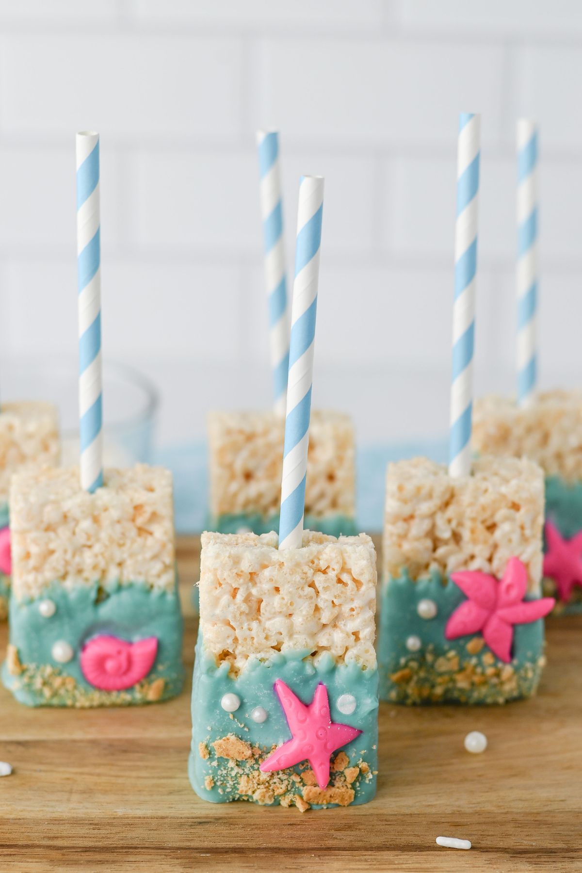 Rice Krispies treats on sticks, dipped in blue coating with pink starfish and seashell decorations, and topped with cookie crumbs to resemble a beach scene.