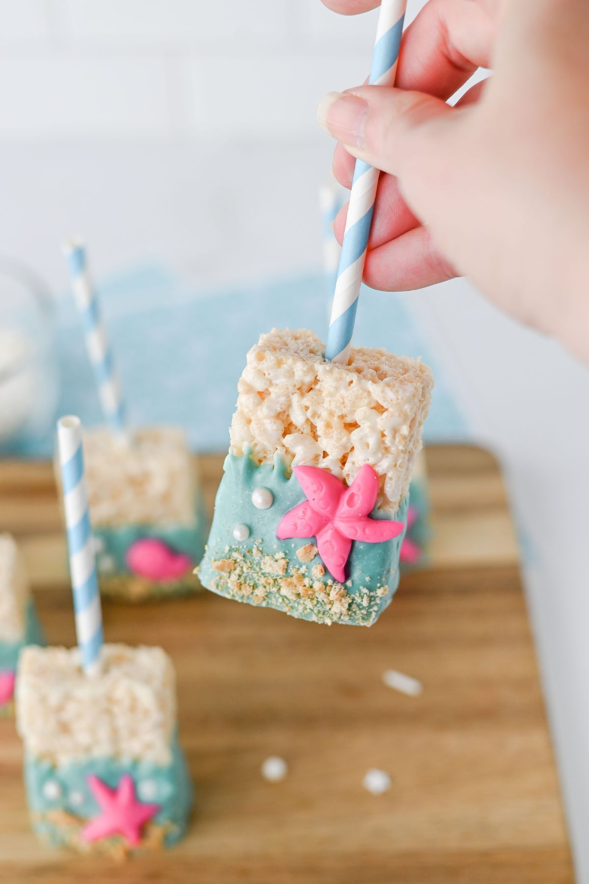 A hand holds a Rice Krispies treat on a stick, dipped in blue coating and decorated with a pink starfish and sprinkles, with more treats on a wooden board below.