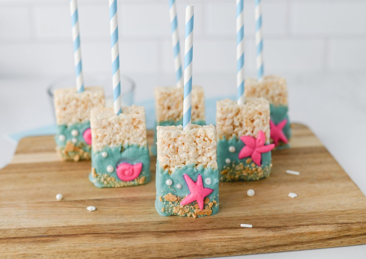 Rice Krispies treats on sticks, decorated with blue icing, pink sea creatures, white dots, and crushed crumbs, arranged on a wooden board.