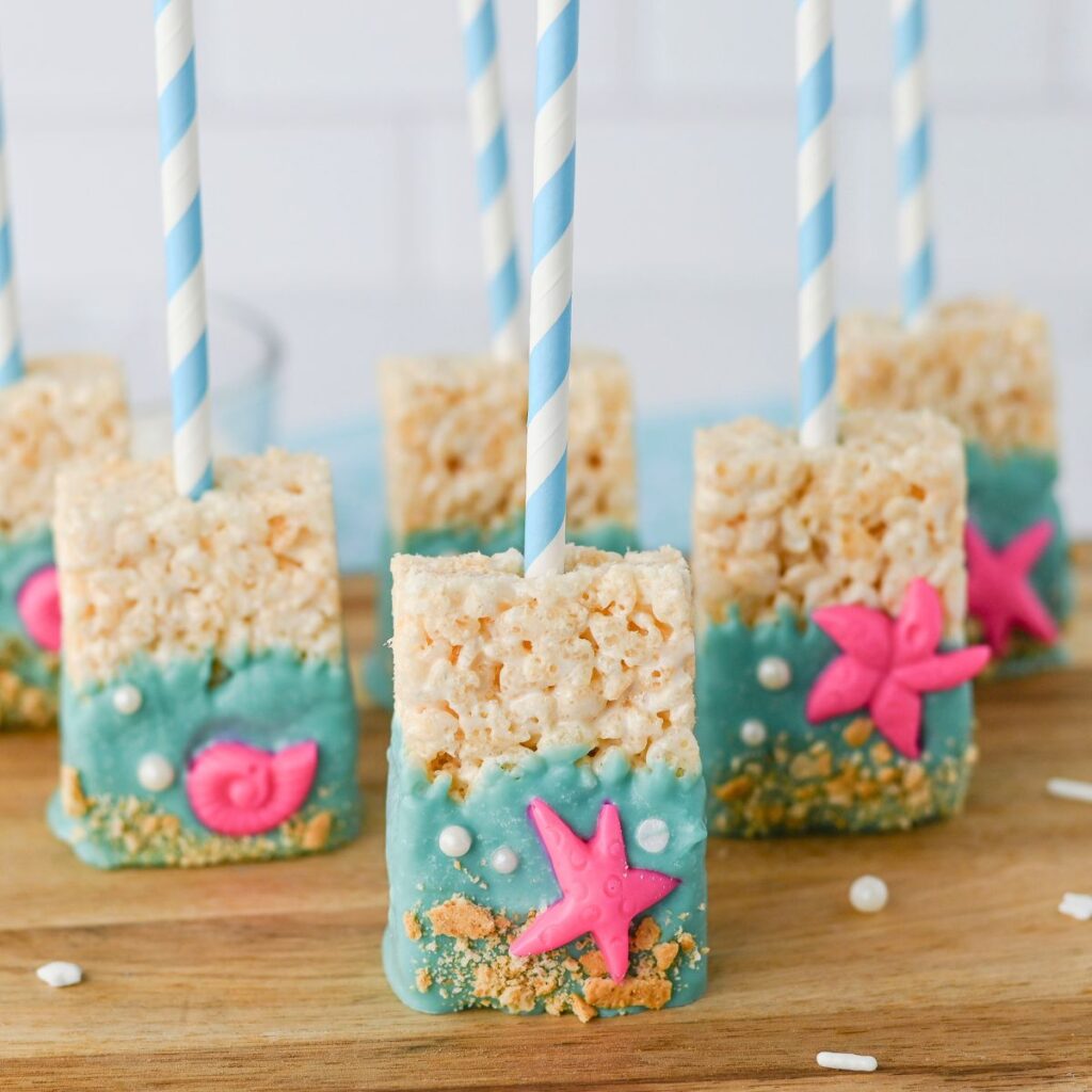 Rice Krispies treats on sticks are dipped in blue candy coating and decorated with pink seashell and starfish shapes, white pearls, and cookie crumbs to resemble an ocean beach scene.