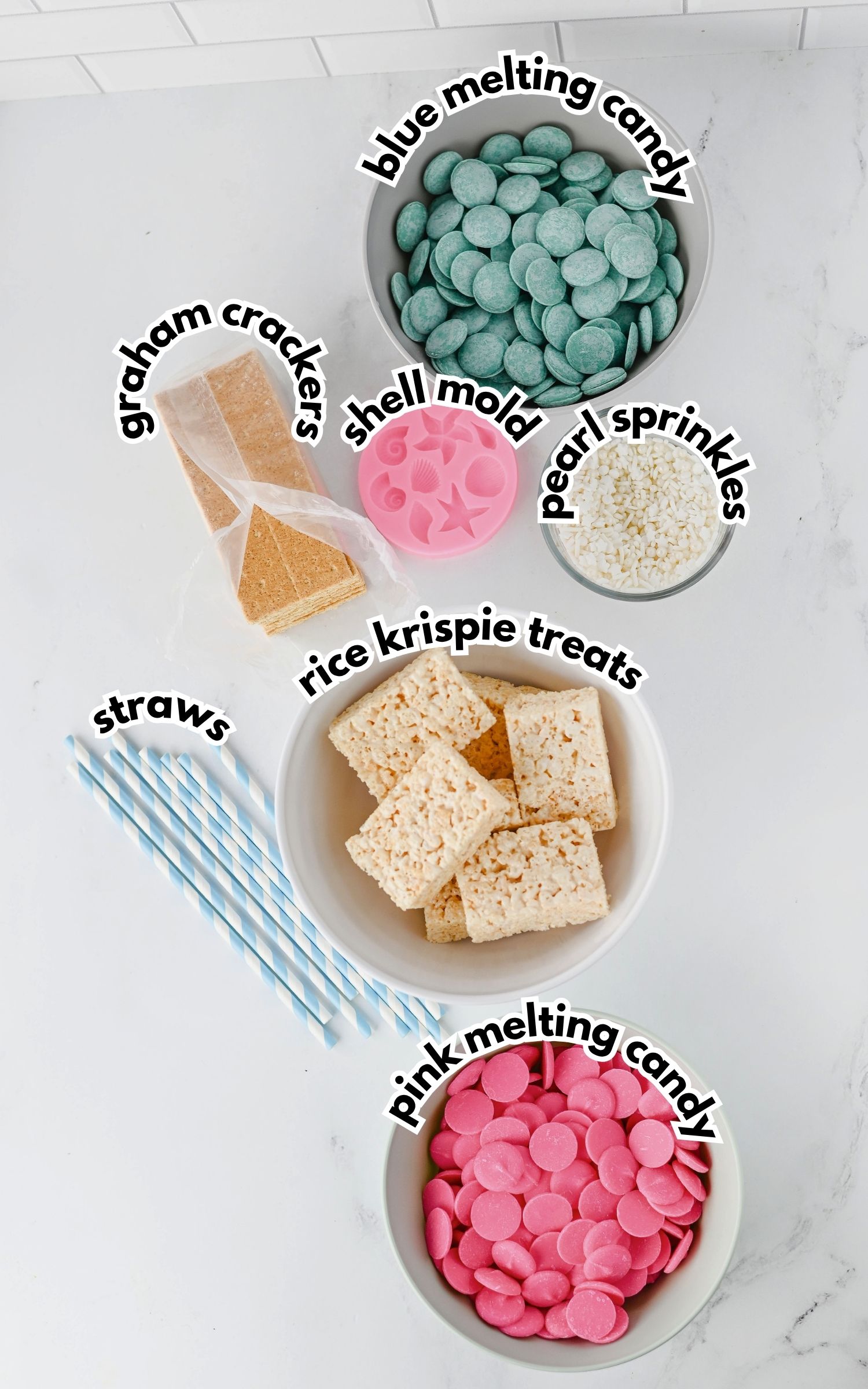 Bowls containing blue melting candy, pink melting candy, pearl sprinkles, graham crackers, rice krispie treats, a shell mold, and striped paper straws arranged on a white surface.