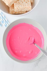 A bowl of bright pink melted candy coating with a spoon inside, next to a bowl of Rice Krispies treats and several blue striped straws.