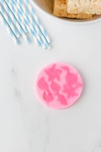 A round pink silicone mold with sea animal shapes, a few blue and white striped straws, and a bowl of Rice Krispies treats on a white surface.