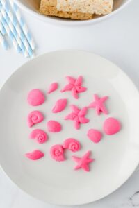 A white plate with small, pink sea-themed candies shaped like shells and starfish; striped blue straws and a bowl of rice treats in the background.