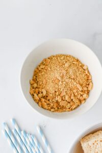 A white bowl filled with crushed graham crackers sits on a white surface next to several blue and white striped paper straws.