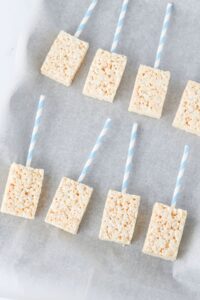Nine rectangular Rice Krispies treats with blue and white striped sticks inserted, arranged on a sheet of parchment paper.