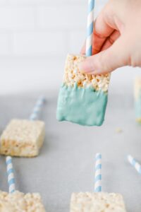 A hand holds a rice krispies treat on a blue striped stick, partially dipped in blue frosting, above a parchment-lined surface with more treats.