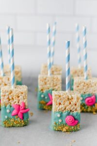 Rice Krispie treats on sticks dipped in blue coating, decorated with pink sea-themed fondant shapes and cookie crumbs, arranged on a light surface with a white tiled background.