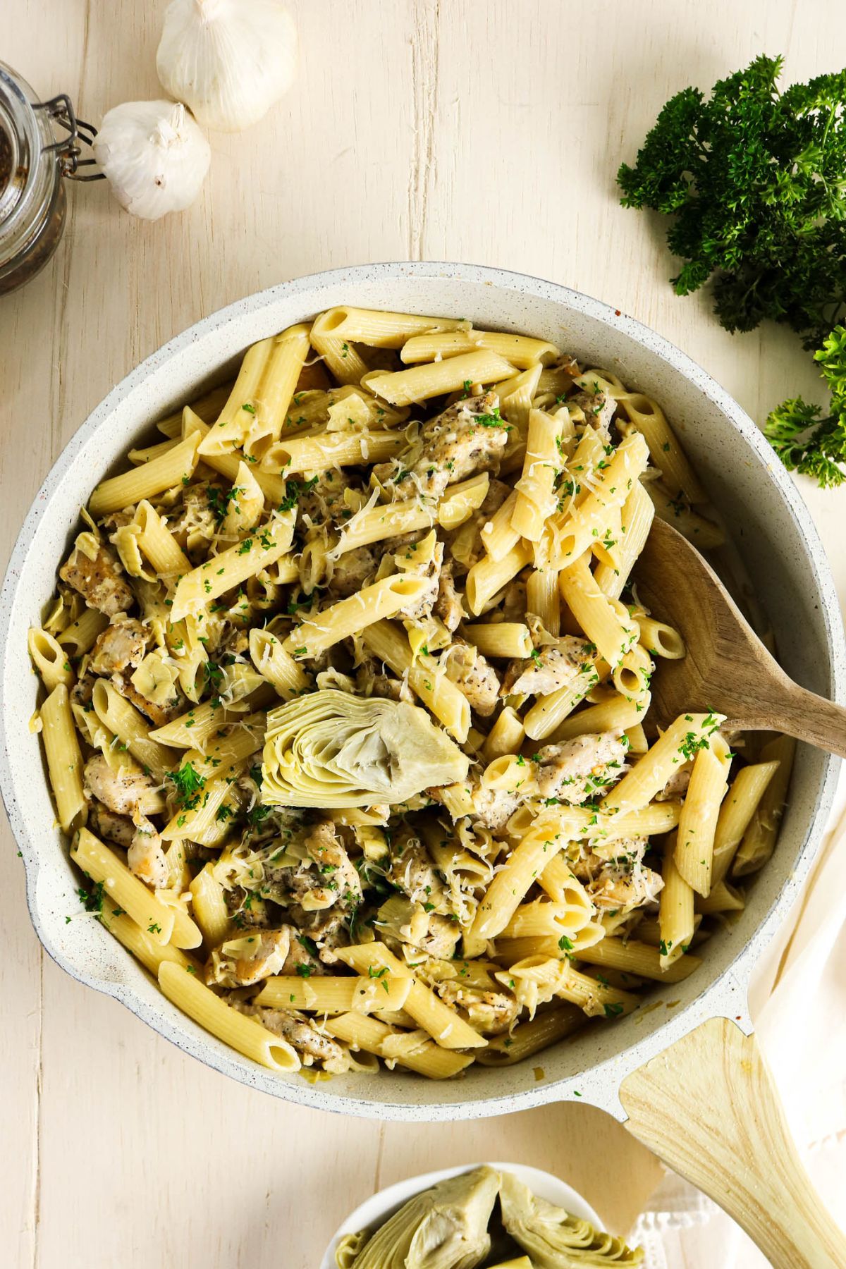 A skillet of penne pasta with artichoke hearts, herbs, and grated cheese, with a wooden spoon and fresh parsley and garlic nearby.