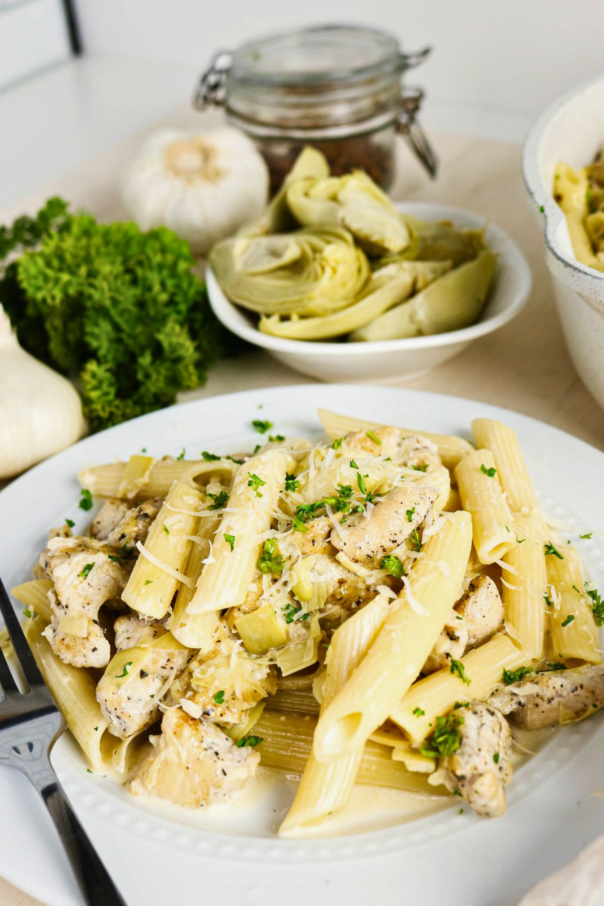A plate of penne pasta with pieces of chicken, artichoke hearts, and parsley, with fresh garlic, parsley, and a bowl of artichoke hearts in the background.