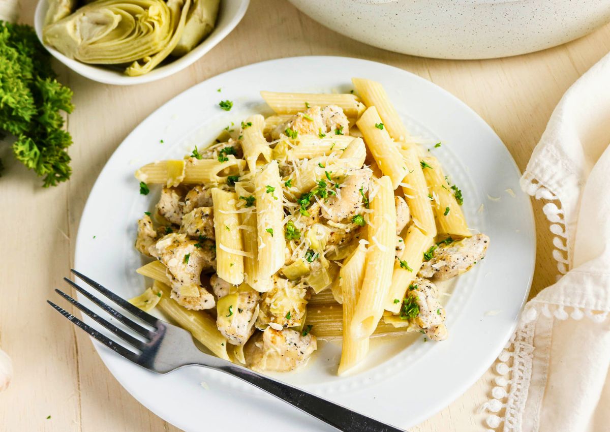 A plate of penne pasta with pieces of chicken and artichokes, garnished with chopped parsley and grated cheese, sits on a white table next to a fork and a bowl of artichoke hearts.