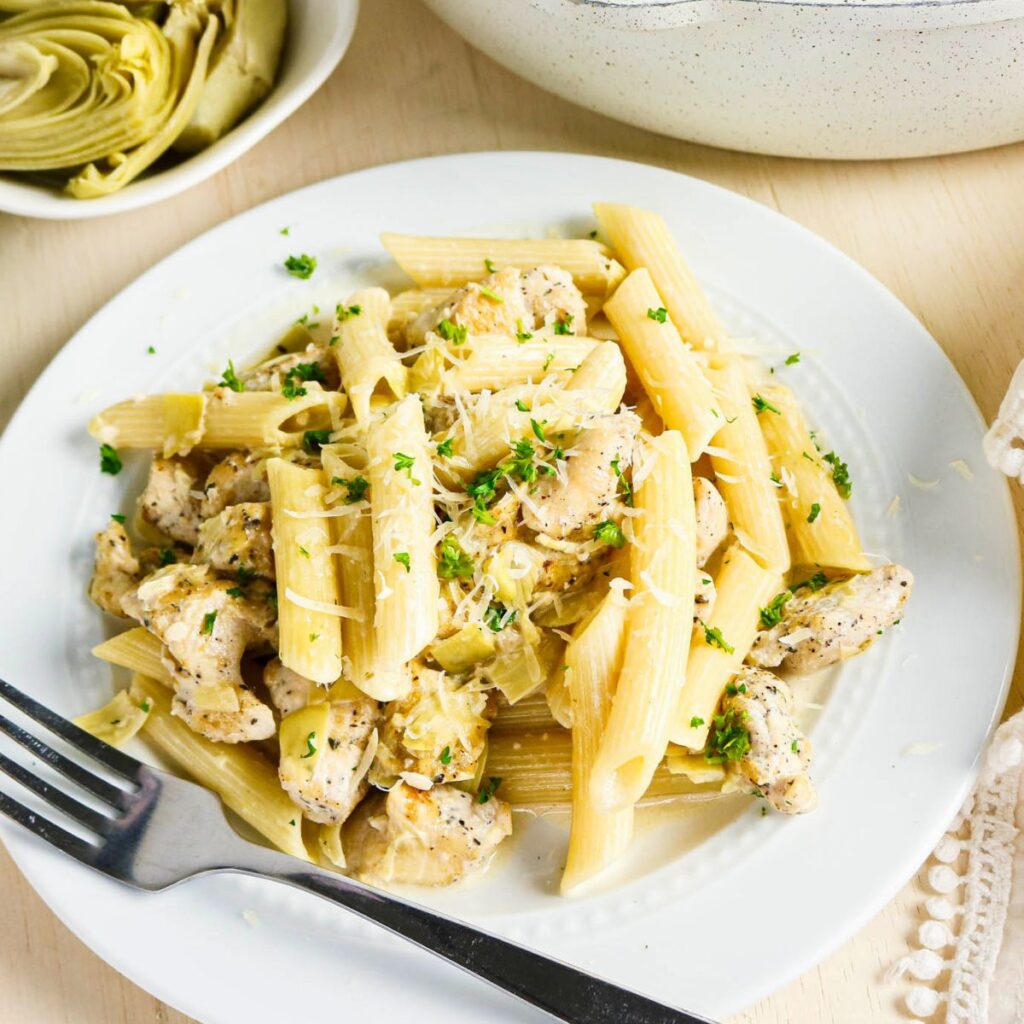 A plate of penne pasta with pieces of chicken, topped with grated cheese and chopped herbs, served with a fork on a white plate. A bowl of artichoke hearts is nearby.