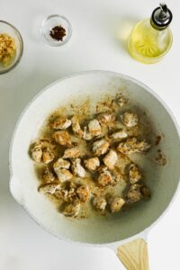 Cubed chicken pieces cooking in a white skillet, with bowls of minced garlic, red pepper flakes, and a bottle of olive oil nearby on a white surface.