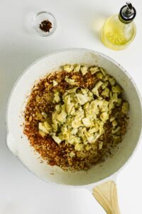 A white skillet with sautéed garlic and chopped artichoke hearts, next to a bottle of oil and a small dish of red pepper flakes on a white surface.