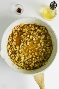 A white skillet with chopped artichokes cooking in liquid, next to a small bowl of spices and a bottle of oil on a white surface.