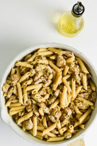 A pan filled with penne pasta and chicken pieces sits on a white surface next to a bottle of olive oil.