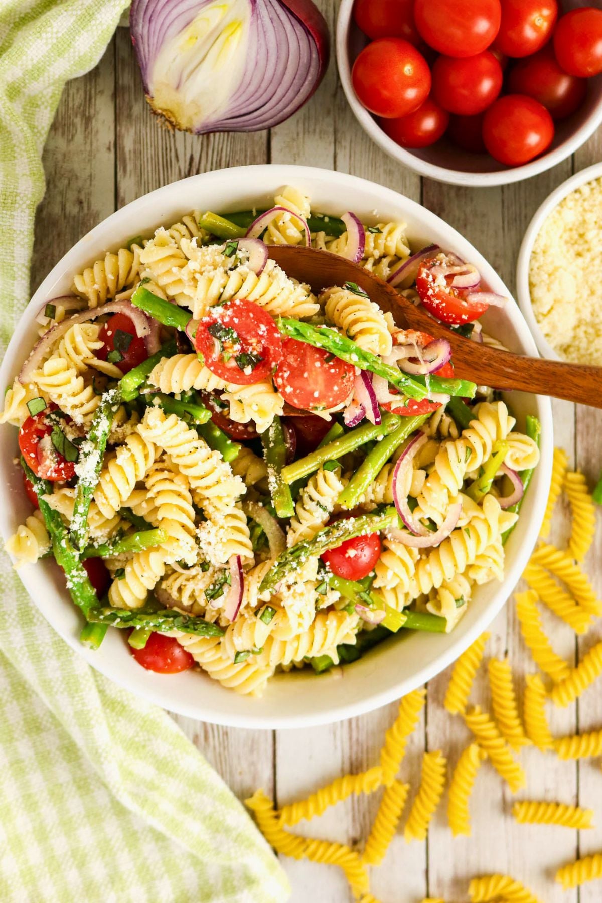 A bowl of rotini pasta salad with cherry tomatoes, asparagus, red onion, grated cheese, and a wooden spoon, surrounded by ingredients on a wooden surface.